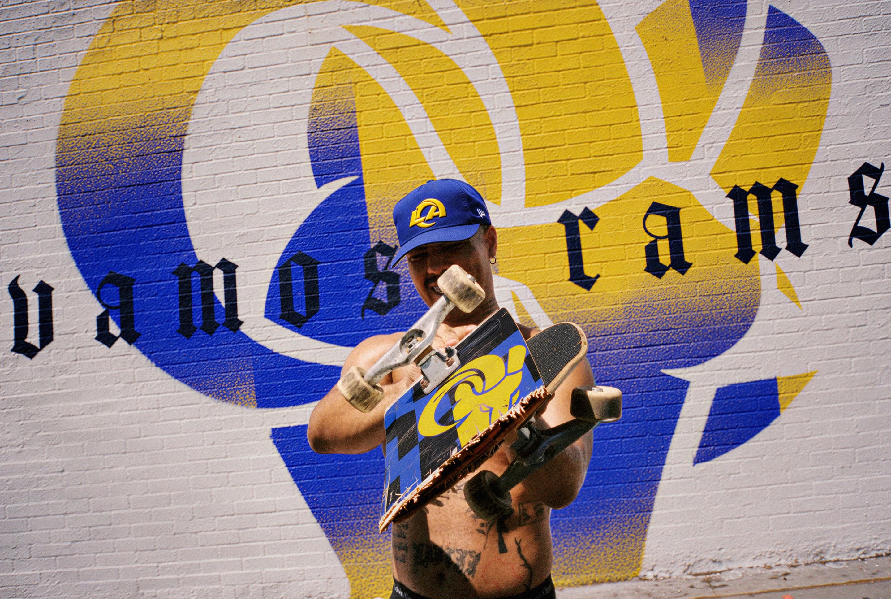 A skater holding his board in front of the Vamos Rams mural