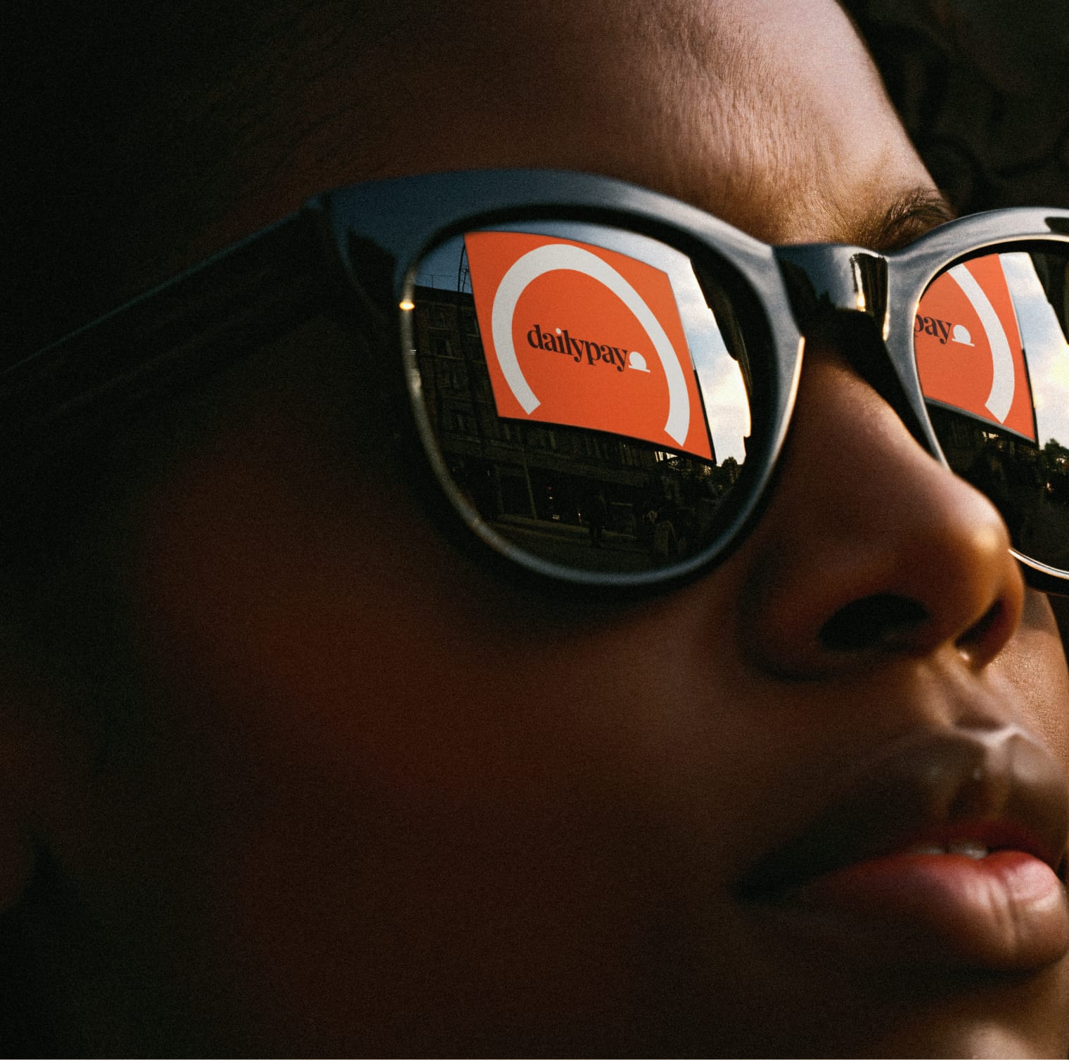 DailyPay brand portrait — a person in sunglasses reflecting a DailyPay billboard at dusk