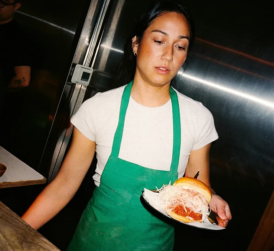 A server in a green apron carrying a plated burger through a restaurant