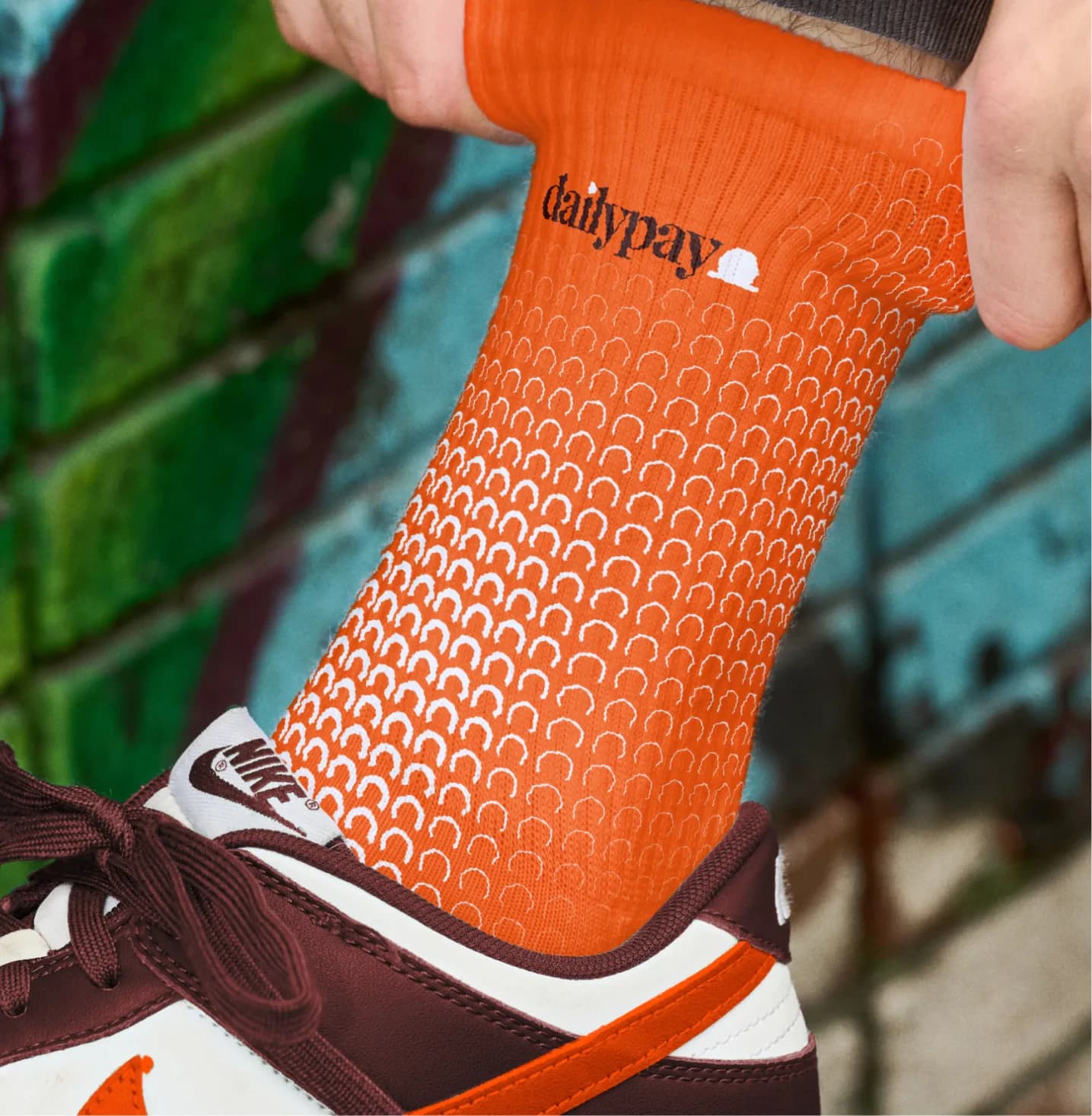 An orange dailypay sock patterned with white arches, above a maroon Nike trainer