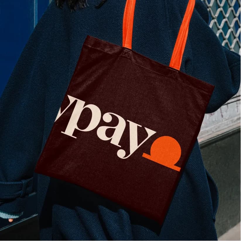 A dark tote bag with orange handles carrying the dailypay wordmark, worn over a blue coat