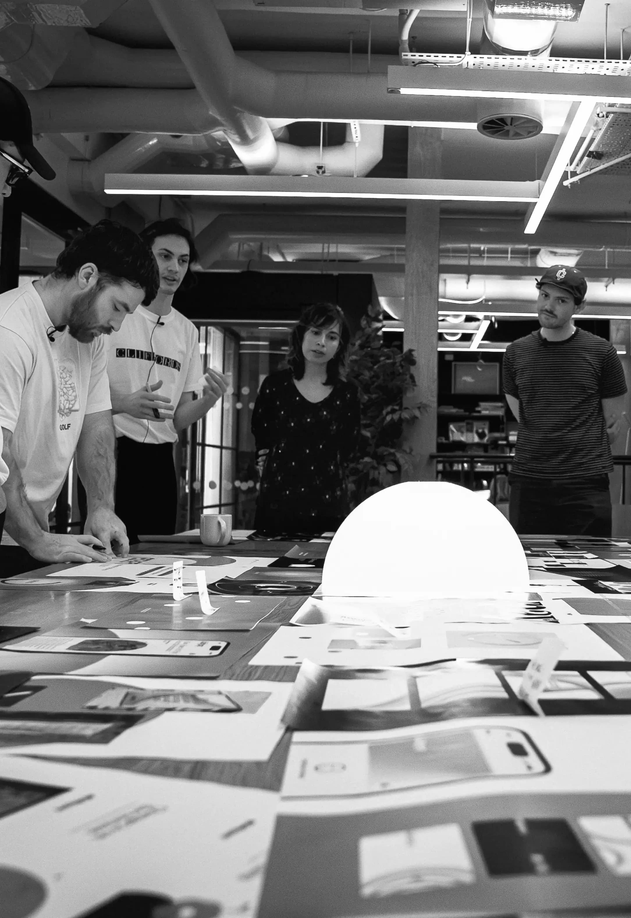 Four people around a table covered in printed layouts, a glowing dome lamp at the centre
